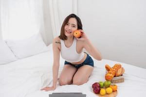 Portrait of white Asian woman with a tattoo sitting on the bed relaxing and playing laptop on weekend There was bread and fruit and red apples on a white bed to eat. holiday concept photo