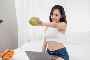Portrait of a white Asian woman with tattoos sitting holding green apples and fruits in a healthy lifestyle. Relaxing in bed and playing on my laptop for the weekend. holiday concept. photo