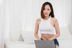 Portrait of a white Asian woman with a tattoo sitting resting on a bed and playing laptop for a working holiday, vacation concept. photo