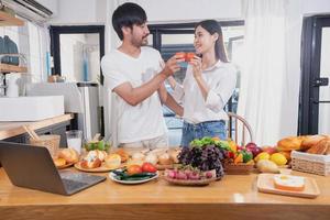 Young Asian couple cooking with fruits and vegetables and using laptop in the kitchen To cook food together within the family happily, family concept. photo