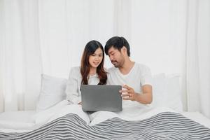 Young happy asian couple using laptop in bed watching movie, romantic time to enhance family relationship, family concept. photo