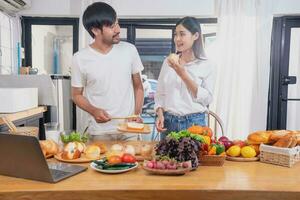 Young Asian couple cooking with fruits and vegetables and using laptop in the kitchen To cook food together within the family happily, family concept. photo