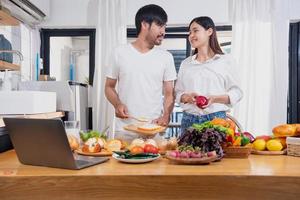 joven asiático Pareja Cocinando con frutas y vegetales y utilizando ordenador portátil en el cocina a cocinar comida juntos dentro el familia felizmente, familia concepto. foto