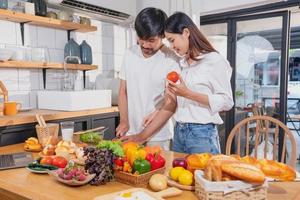 Young Asian couple cooking with fruits and vegetables and using laptop in the kitchen To cook food together within the family happily, family concept. photo