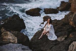 un mujer se sienta en un acantilado en un blanco vestir por el Oceano nublado clima inalterado foto