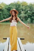 A young woman in a hippie look and eco-dress travels in nature by the lake wearing a hat and yellow pants in the fall sunset photo