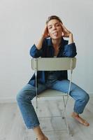 A young woman sitting in a chair at home smiling with teeth with a short haircut in jeans and a denim shirt on a white background. Girl natural poses with no filters photo