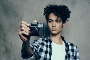 joven hombre con un cámara en mano y en un tartán camiseta en un gris antecedentes adentro fotógrafo foto