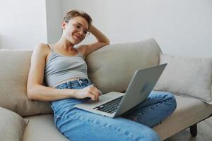 Teenage girl freelancer with laptop sitting on couch at home smiling in home clothes and glasses with short haircut, lifestyle with no filters, free copy space photo