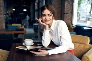 happy woman in shirt and red skirt sits at a table in a cafe with notepad and pen in hand photo