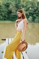 A young woman smiling in an image of a hippie and eco-dress dancing in nature by the lake wearing a hat and yellow pants in the summer sunset photo