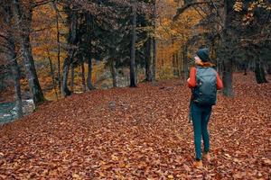 woman with backpack hiking travel in autumn park tall trees river fallen leaves photo