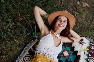 un mujer en Respetuoso del medio ambiente ropa en un hippie Mira es acostado en un de colores tartán sonriente y mirando a un otoño puesta de sol en naturaleza en el parque. estilo de vida en un viaje de armonía con el mundo foto