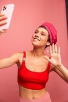 A young woman blogger with colored pink hair and a short haircut takes a picture of herself on the phone and broadcasts a smile in stylish clothes and a hat on a pink background monochrome style photo