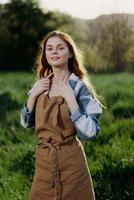 A woman gardener in an apron stands in a field of green grass outdoors, smiling on a summer day into a sunset photo