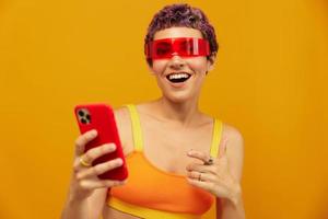 Woman blogger surprise in unusual millennial glasses taking selfies on her phone sporting brightly colored clothes against an orange studio background, free space photo