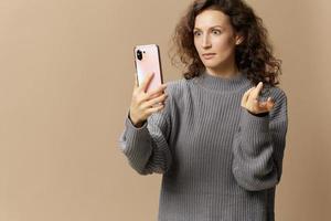 Shocked irritated curly lady in gray casual sweater do video call with phone get bad news posing isolated on over beige background. Social media, network, distance communication concept. Copy space photo