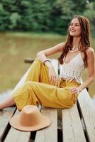 A hippie woman sits on a bridge by a lake on a nature trip and smiles happily in eco-clothing. photo