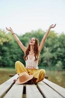 A hippie woman sits on a bridge by a lake with her hands up in the air while traveling in nature and smiling in eco-clothing. Relaxed lifestyle photo