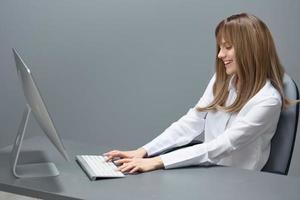 Happy pretty blonde businesswoman worker freelancer using desktop computer chatting with clients in gray modern office of support service. Remote Job, Technology Career Profession Concept. Copy space photo