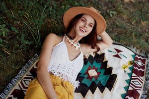 un mujer en Respetuoso del medio ambiente ropa en un hippie Mira es acostado en un de colores tartán sonriente y mirando a un otoño puesta de sol en naturaleza en el parque. estilo de vida en un viaje de armonía con el mundo foto