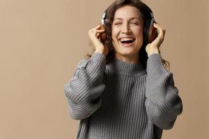 Joyful smiling curly beautiful female in gray casual sweater wear headphones enjoy cool playlist posing isolated on over beige pastel background. Sound streaming platform concept. Copy space photo