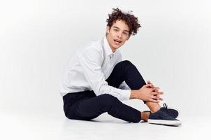 curly-haired guy in shirt and trousers and sneakers sits on the floor indoors side view photo