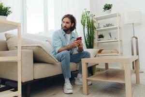 A man with a beard sits on the couch during the day and smiles at home and looks at his phone relaxing on his day off, life online on his phone texting with friends photo
