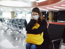 cansado mujer en médico máscara con equipaje aeropuerto esperando vuelo retrasar foto