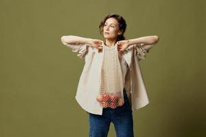 Serious cute curly beautiful lady in linen casual shirt pulls string bag with fruit near chest posing isolated on over olive green pastel background. Eco-friendly Natural products concept. Copy space photo