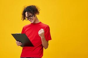 Joyful happy myopic young student man in red t-shirt funny eyewear holds tablet folder with study notes posing isolated on over yellow studio background. Free place for ad. Education College concept photo
