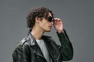 Side of view portrait of stylish tanned curly man leather jacket wear glasses posing isolated on over gray background. Cool fashion offer. Huge Seasonal Sale New Collection concept. Copy space for ad photo