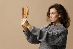 Pensive adorable curly beautiful female in gray casual sweater find perfect hairbrush comb posing isolated on over beige pastel background. Problematic unruly damaged hair concept. Copy space photo