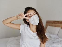 woman in medical mask holds two fingers near her eyes and tilts her head to the side indoors photo