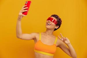 Woman blogger smiling in unusual millennial glasses taking selfies on her phone in sportswear against an orange studio backdrop, free space photo