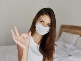 woman in medical mask sitting on bed in hotel positive hand gesture photo