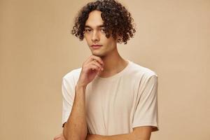 Pensive funny awesome tanned curly man in basic t-shirt can not make decision posing isolated on beige pastel background. Fashion New Collection offer. People and Emotions concept. Free place for ad photo