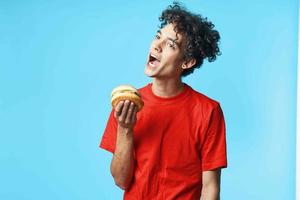 alegre chico con Rizado pelo en un rojo camiseta con un hamburguesa en su manos rápido comida dieta foto