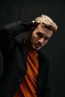 blond guy in an orange sweater and jacket on a dark background holds his hand behind his head close-up cropped view photo