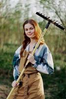 Woman beautifully smiling farmer in work clothes and apron working outdoors in nature and holding a rake to gather grass photo