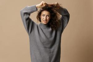 Tormented curly beautiful female in gray casual sweater with hairbrush comb pulls tangled hair posing isolated on over beige pastel background. Problematic unruly damaged hair concept. Copy space photo