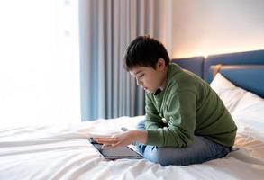 Kid boy using tablet playing game on internet with friend, Homeschooling Kid doing homework online by digital pad at home,Child sitting on bed relaxing,watching cartoon or talking online with friend photo