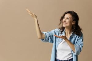 Cheerful happy curly beautiful lady in jeans casual shirt point both hands looks up at free place for ad posing isolated on over beige pastel background. People Lifestyle emotions concept. Copy space photo