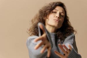 Friendly flirted curly beautiful female in gray warm casual sweater pulls hands send kiss at camera posing isolated on beige pastel background. Fashion Sale offer. People emotions concept. Copy space photo