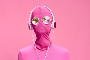 Art portrait of a woman wearing a pink full-face burglar mask with glowing round glasses wearing pink clothes with pink headphones on a pink background looking into the camera photo