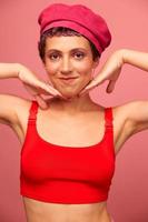 joven atlético mujer con un corto Corte de pelo y púrpura pelo en un rojo parte superior y un rosado sombrero con un atlético figura sonrisas y muecas mirando a el cámara en un rosado antecedentes foto