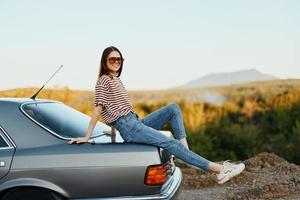 un mujer con un coche detenido en el la carretera a descanso en el viaje elevado su brazos y piernas desde felicidad y un hermosa paisaje foto