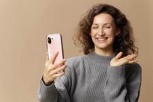 Friendly curly beautiful female in gray casual sweater doing video call with phone posing isolated on over beige pastel background. Social media, network, distance communication concept. Copy space photo