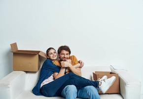 man and woman sitting on a white sofa in a room with boxes of things tools moving photo