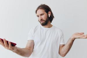 A man with a beard blogger in a white T-shirt with a phone and wireless headphones looks into the phone and spreads his hands to the side on a white background isolated copy space photo